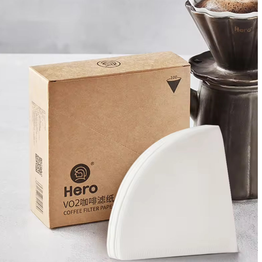 Stack of white cone-shaped coffee filter papers with a brown 'Hero VO2 coffee filter paper' box and a dark ceramic dripper, for LAMOSE.