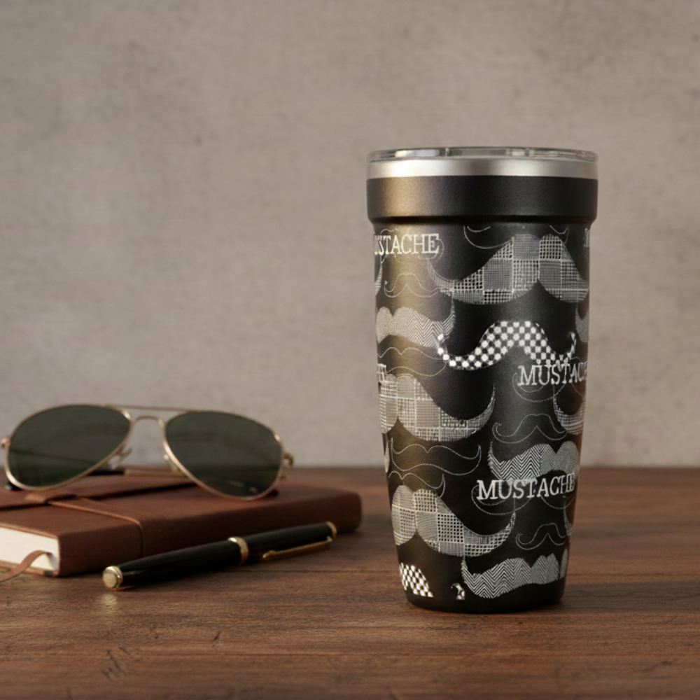 Black 16oz travel tumbler featuring white illustrated mustache patterns standing on a wooden desk next to gold sunglasses, a black pen, and a brown notebook.