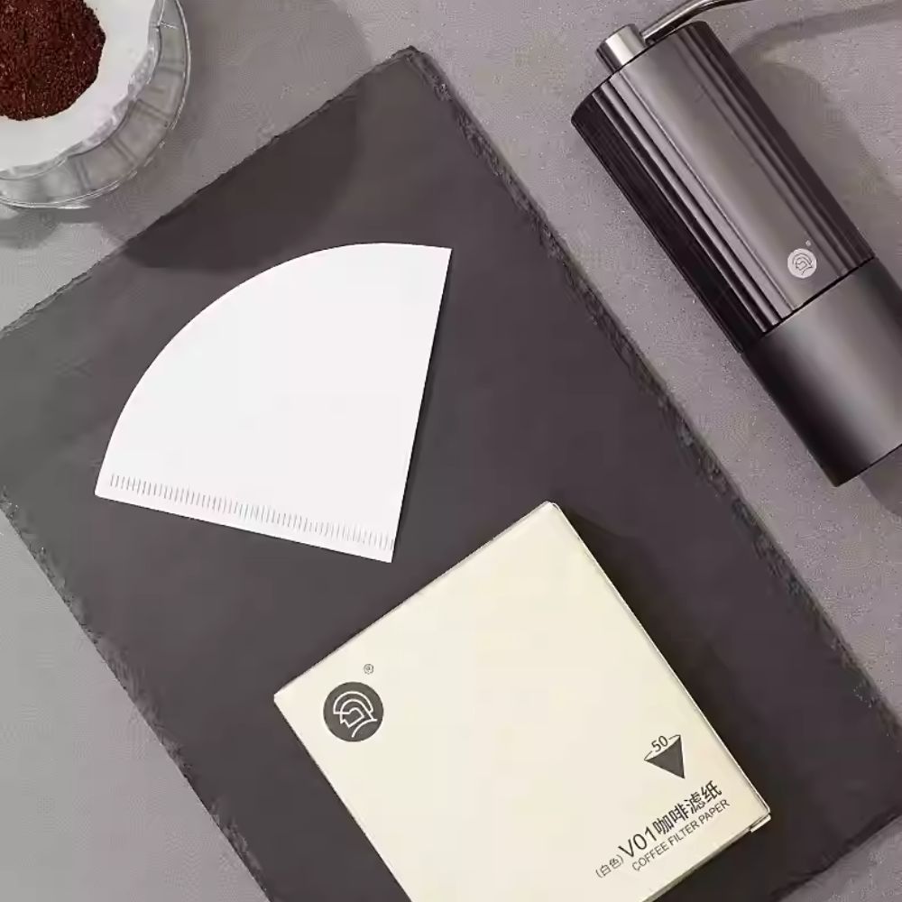White V01 cone coffee filter paper and its cream box with 50 filters, shown from above with a dark grey grinder on a slate surface. LAMOSE.