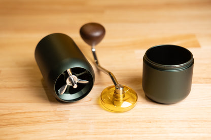LAMOSE zeroHero Z7 manual grinder in dark green, disassembled on wood. Metal burrs, wooden handle, yellow dial, and catch cup shown.