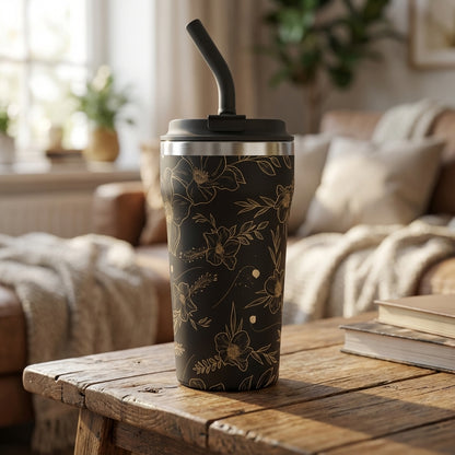 Close up of black matte 16 oz tumbler featuring delicate gold line art of flowers and leaves, standing on a dark wooden surface.
