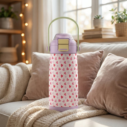 Stainless steel kids water bottle with pink heart pattern and purple lid resting on a cozy sofa
