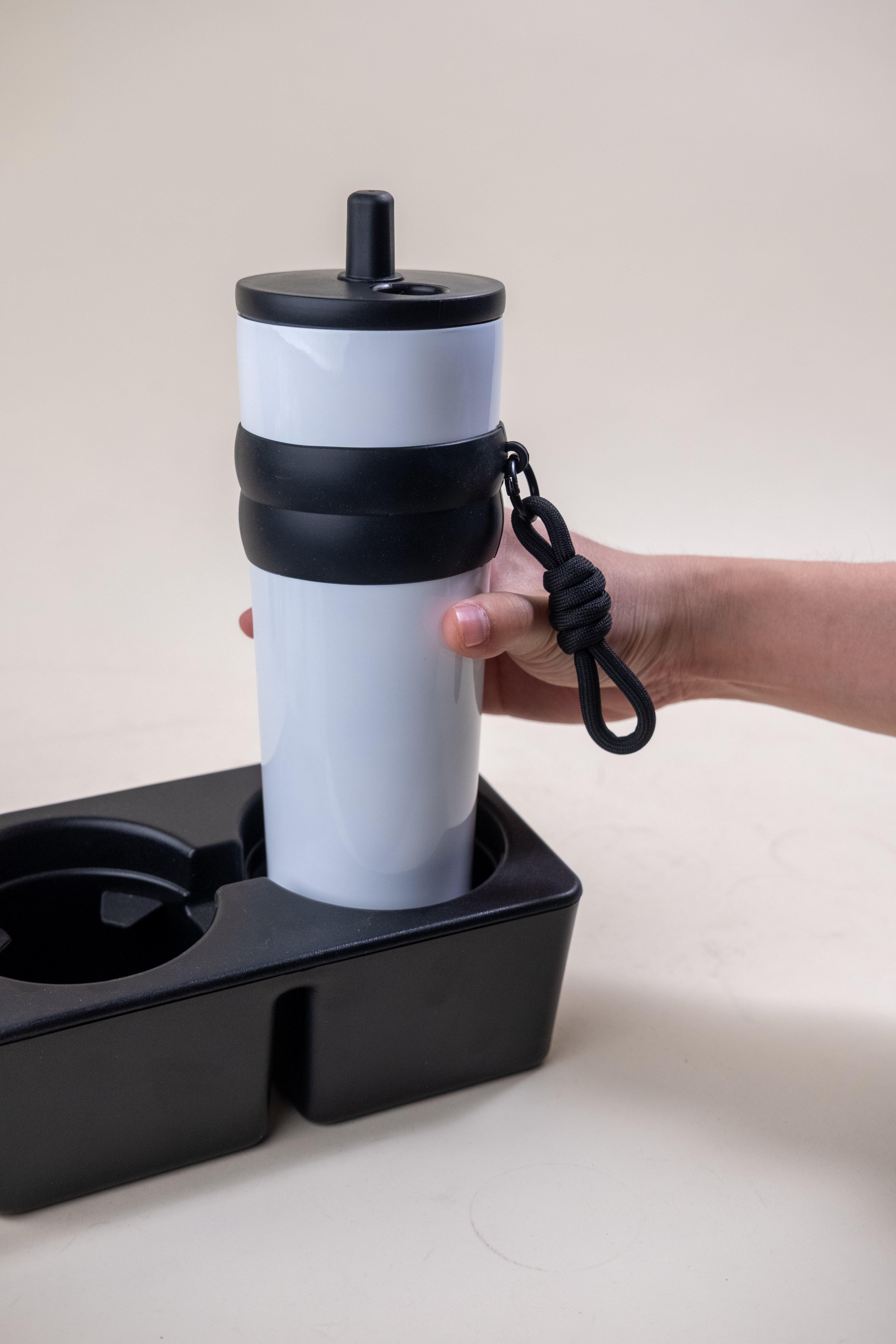 Hand holding a white and black straw tumbler by a sturdy rope, illustrating the tumbler's portability and convenience, perfect for travel or daily commuting. 