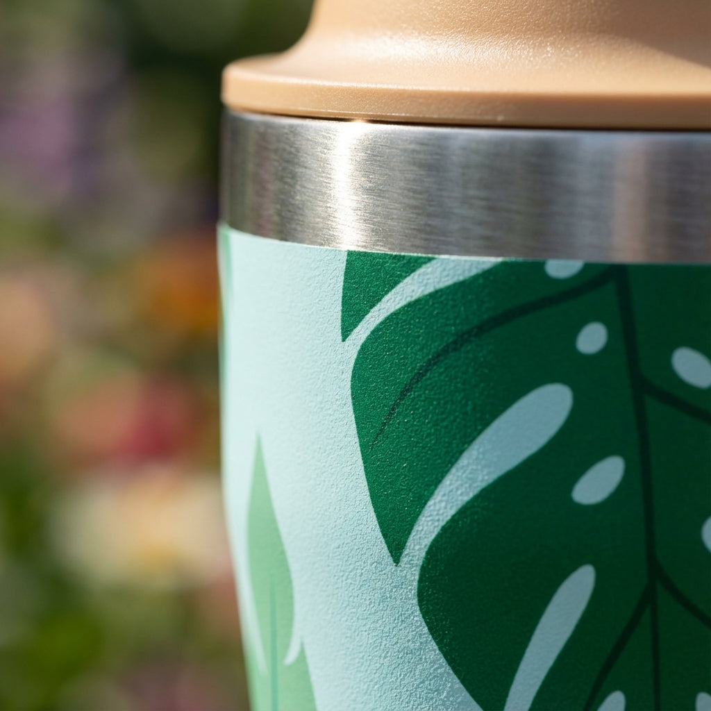 Detail shot - LAMOSE Bow 16 oz light blue tumbler with tropical monstera leaf and pink flower pattern standing in sunlight.