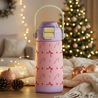 Pink LAMOSE Goose 14 oz kids water bottle with red hearts, bows, and candy cane pattern, lavender silicone boot, and green handle, set against a blurry Christmas tree background.