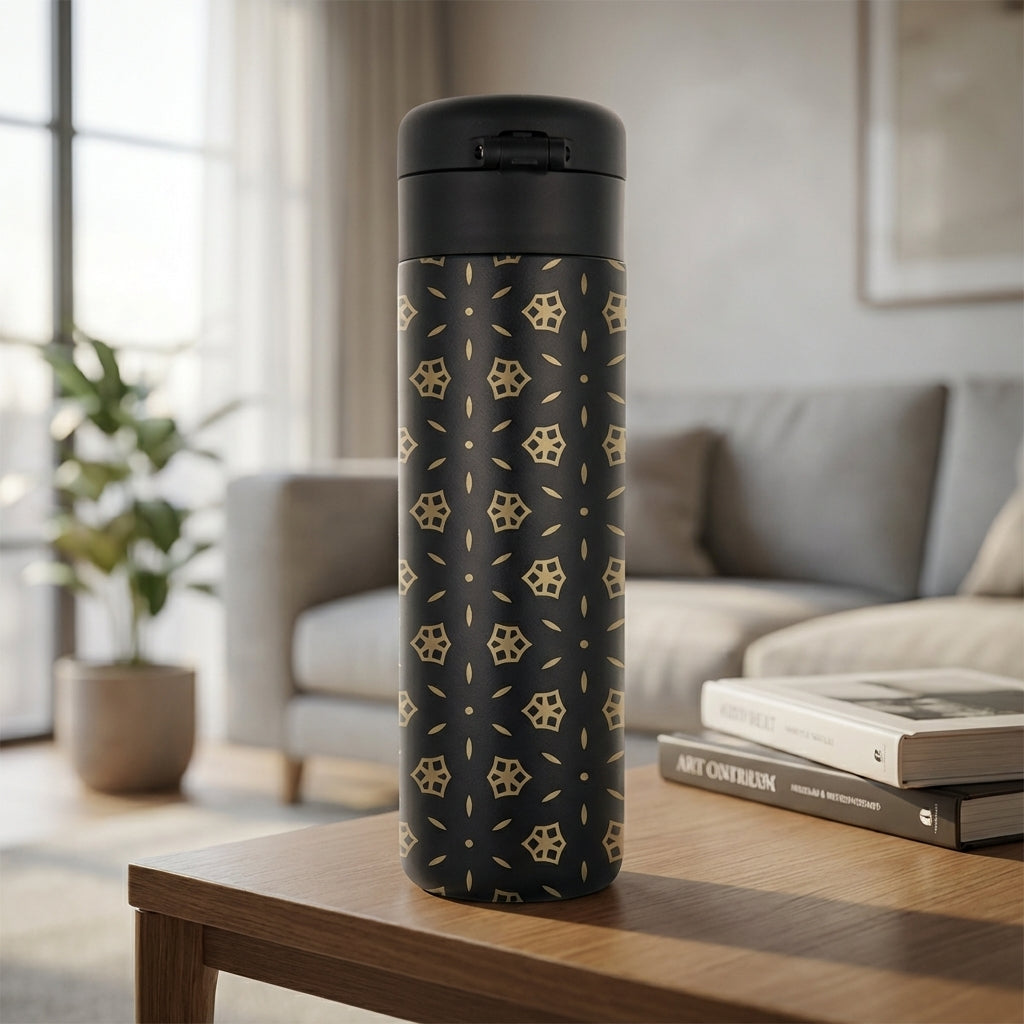 Matte black stainless steel travel tumbler featuring a gold geometric circle and square pattern, sitting on a wooden table in natural light.