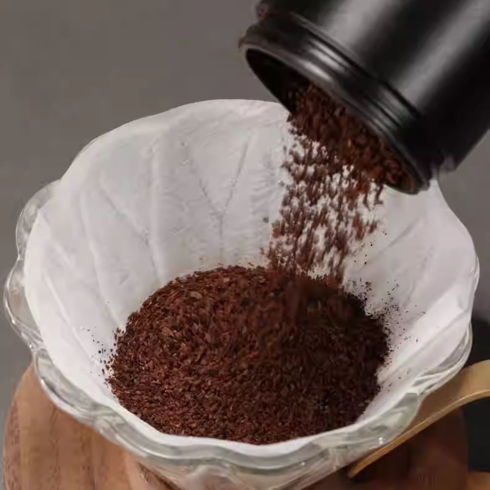 White LAMOSE zeroHero V01 pour-over paper filter in a glass dripper as coffee grounds are poured in. Top-down view.