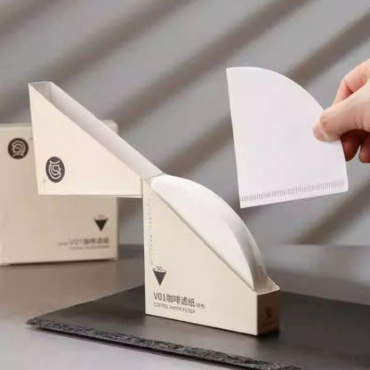 Hand pulling a white V01 conical coffee paper filter from an open light beige zeroHero dispenser box, offering 50 filters. LAMOSE.