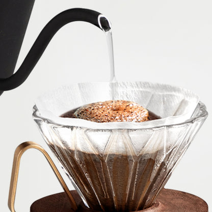 LAMOSE clear glass prisma coffee dripper with gold handle on walnut base, brewing pour-over coffee from a black gooseneck kettle. Close-up.